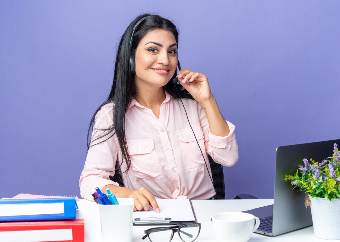 young-beautiful-woman-casual-clothes-wearing-headset-with-microphone-looking-confident-smiling-sitting-table-with-laptop-blue-background-working-office_141793-127770